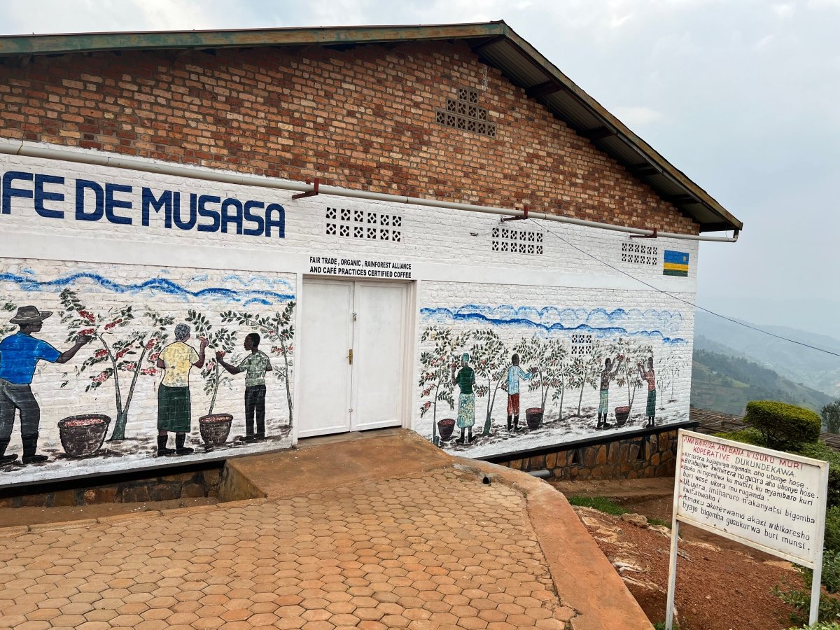 Mural en la fachada de la estación de lavado Musasa en Ruanda, con ilustraciones de caficultores recolectando cerezas. Edificio de ladrillo con certificaciones Fair Trade, Rainforest Alliance y prácticas sostenibles. Reflejo del trabajo comunitario detrás del café de especialidad que Artisan Coffee tuesta en Málaga para espresso y filtrado.
