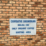 Cartel sobre una pared de ladrillo que indica "Cooperative Dukundekawa Mbilima CWS - Only Organic Coffee Sorting Area", en la estación de café Mbilima, Gakenke, Ruanda. Esta imagen representa el compromiso con el procesamiento ecológico y ético del café de especialidad en origen. Artisan Coffee, tu tostador de café en Málaga, trabaja con cafés orgánicos de cooperativas sostenibles.