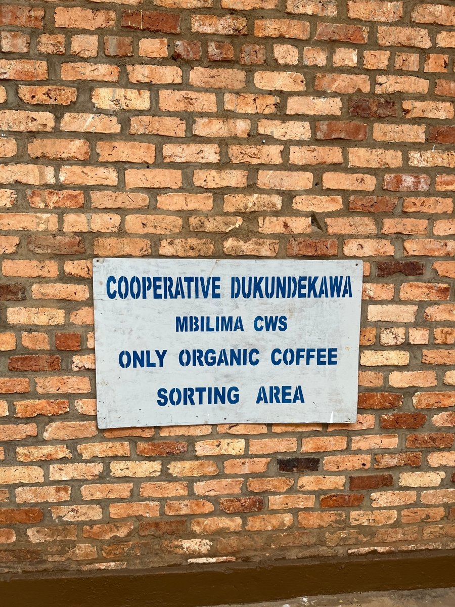 Cartel sobre una pared de ladrillo que indica "Cooperative Dukundekawa Mbilima CWS - Only Organic Coffee Sorting Area", en la estación de café Mbilima, Gakenke, Ruanda. Esta imagen representa el compromiso con el procesamiento ecológico y ético del café de especialidad en origen. Artisan Coffee, tu tostador de café en Málaga, trabaja con cafés orgánicos de cooperativas sostenibles.